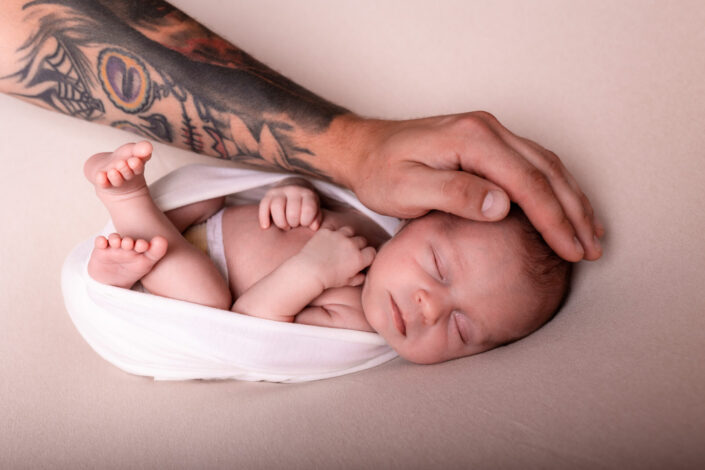 Zdjęciowa sesja noworodkowa w studiu Olsztyn. Mała córeczka a duża dłoń taty. Porównanie małej kruszynki do dużej dłoni taty. Mocny tatuaż a małe bezbronne dziecko.