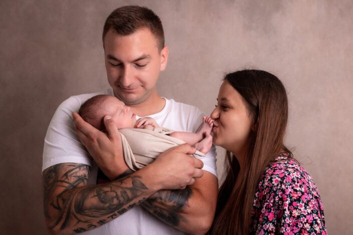 Noworodkowa sesja w studiu Olsztyn Jaroty Bartąska. Zdjęcie noworodka z rodzicami. Ujęcie rodzinne. Zdjęcie rodzinne. Rodzinna sesja zdjęciowa. Mama całuje stópki córeczki.